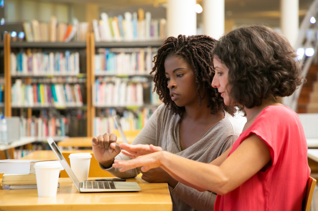 focused-women-talking-pointing-laptop-library_74855-1922